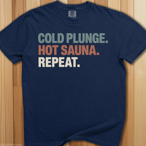 Navy blue t-shirt with 'Cold Plunge. Hot Sauna. Repeat.' text, wooden bucket, and broom on a wooden background.