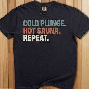Black t-shirt with colorful text 'Cold Plunge. Hot Sauna. Repeat.' on a wooden surface